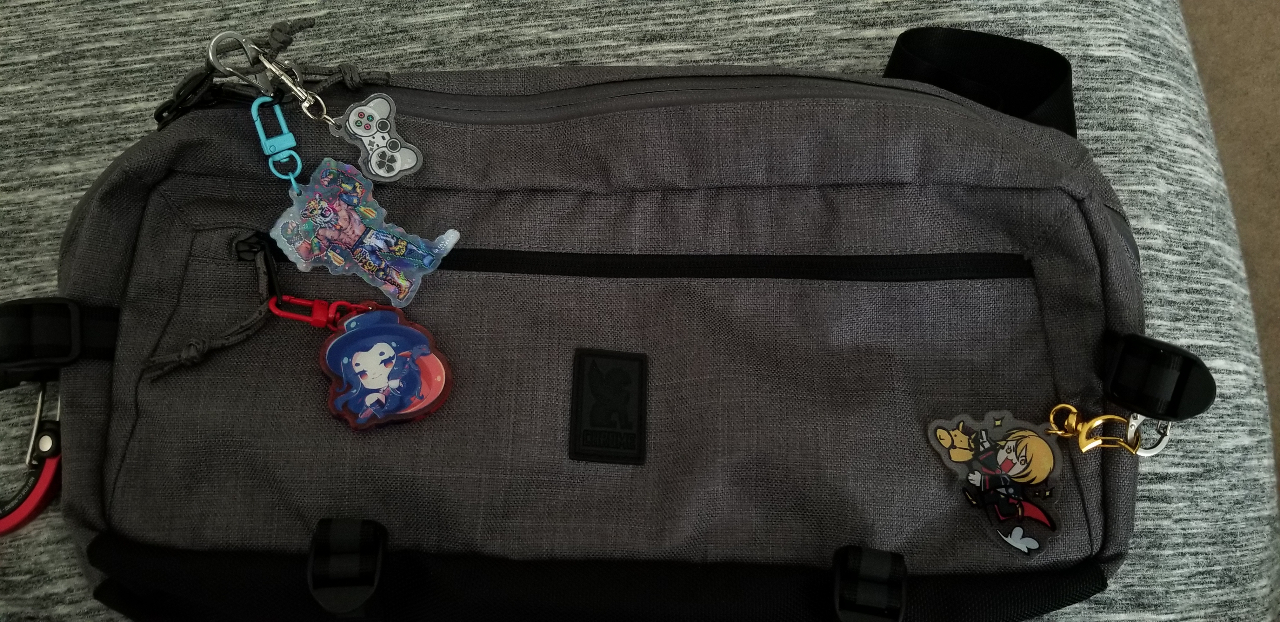 A long, gray sling bag with acrylic charms on its zippers and loop.