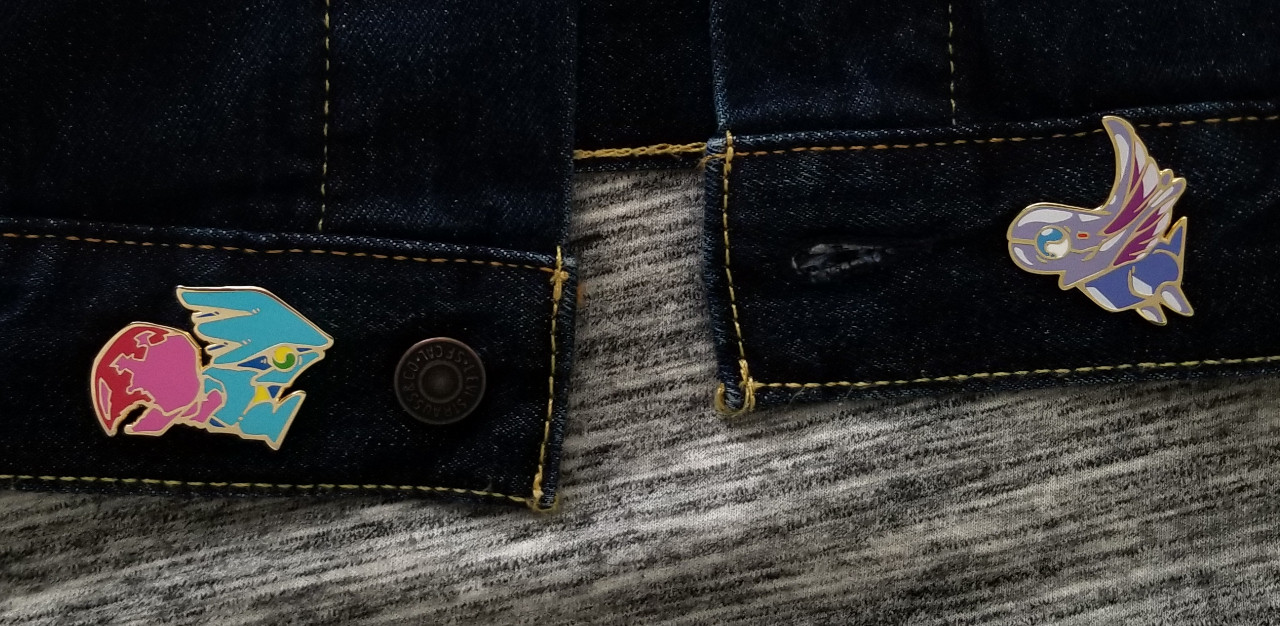 Close up the bottom half of a denim jacket showing off two enamel pins.