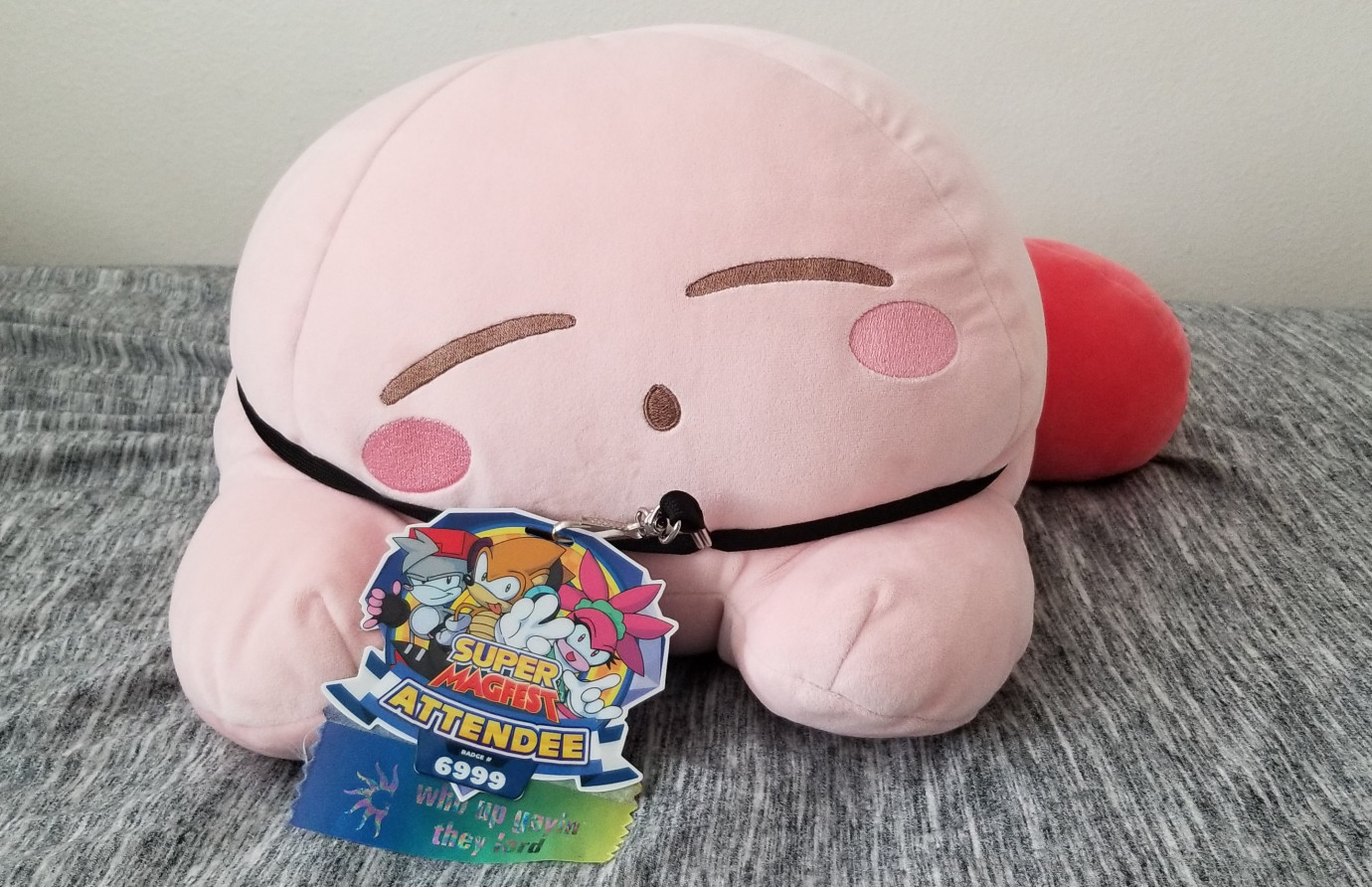 A sleepy Kirby plushie with a lanyard around its body that's holding a MAGfest Attendee badge, #6999, with a ribbon that says "Who's up gaying up they lord?"