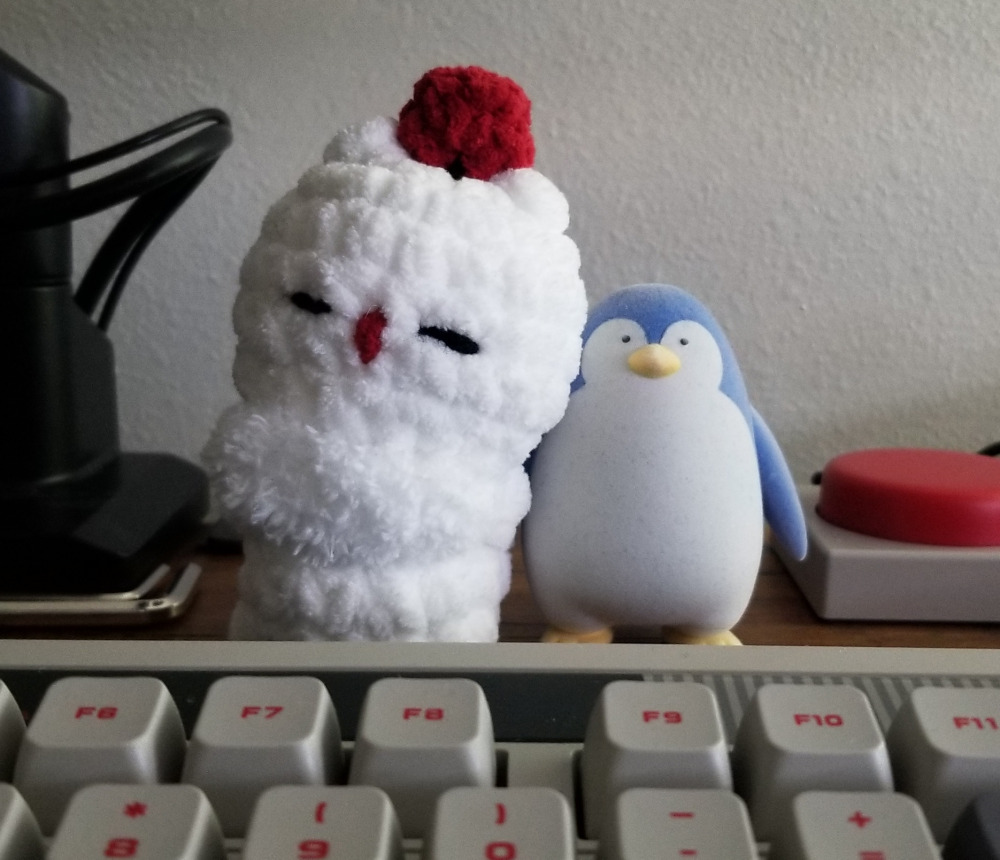 A crochet plush of a Moogle from Final Fantasy. Leaning against a penguin statuette.