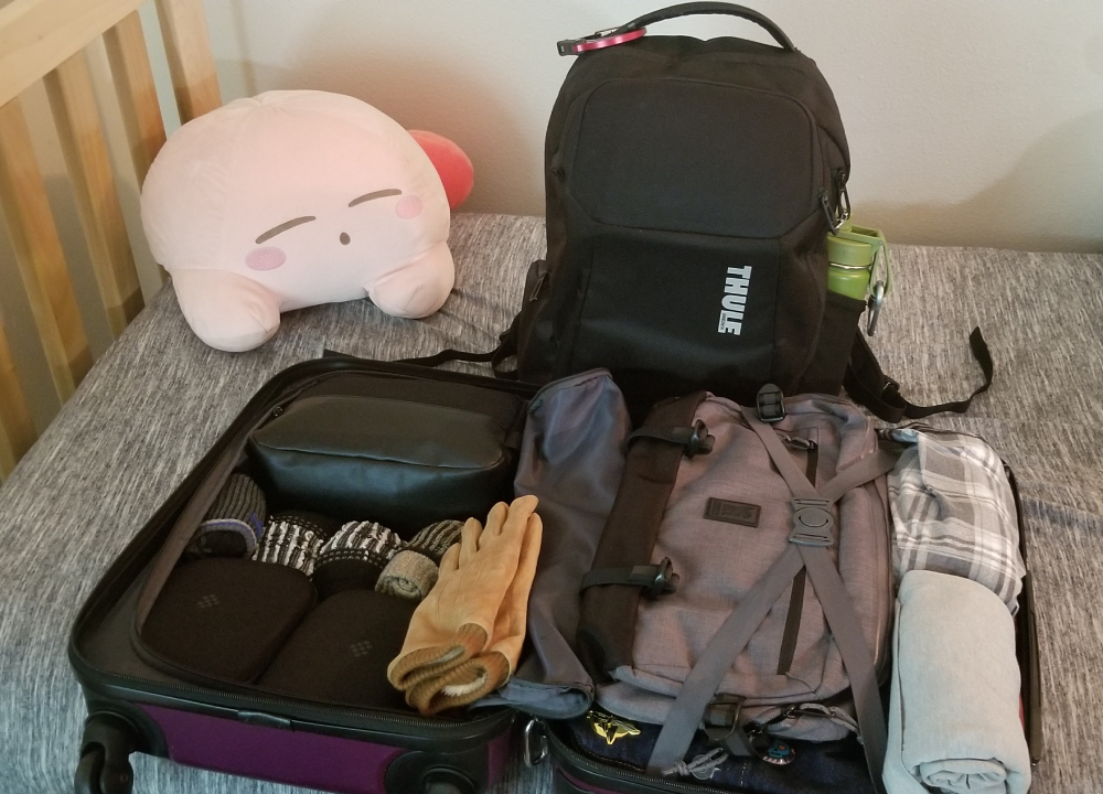 Photo of luggage case open, filled with clothes, and a backpack on a bed with a Kirby plush in the background sleeping.